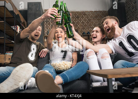 Glückliche junge Freunde Bierflaschen klirren beim Sitzen auf dem Sofa Stockfoto