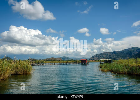 Eine Hütte auf das Ende eines hölzernen Pier in einer Entfernung auf einen See mit grünem Schilf unter blauem Himmel mit flauschigen weissen Wolken Stockfoto