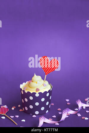 Vanilla Cupcake mit kleinen dekorativen Herzen und roten Herzen Topper, in violett Backpapier Tasse, gegen violette Hintergrund Cupcake für Valentinstag w Stockfoto