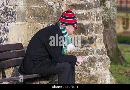 Mann in einem Hut und Schal draußen sitzen auf einer Bank an einem kalten Wintertag in Großbritannien. Stockfoto