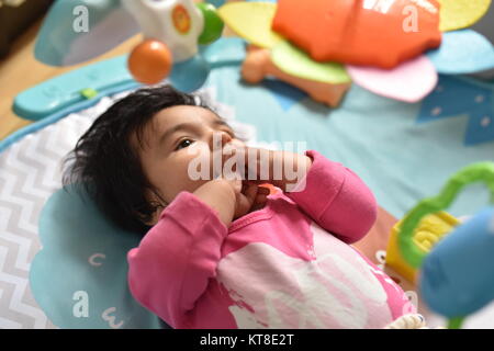 Baby Mädchen mit den Händen auf den Mund beobachtet aufmerksam ihre farbenfrohen Spielzeuge aus einem spielteppich Stockfoto