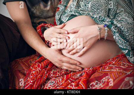 Zugeschnittenes Bild eines schönen schwangeren Frau und ihrem schönen Mann ihr Bauch sich umarmen. In der traditionellen indischen Outfit shalwar kamiz gekleidet. Stockfoto
