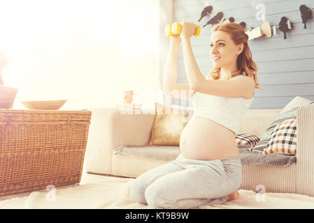 Morgen Übungen. Fröhliche schöne schwangere Frau Training mit Hanteln und kümmert sich um ihre Gesundheit zu Hause Stockfoto