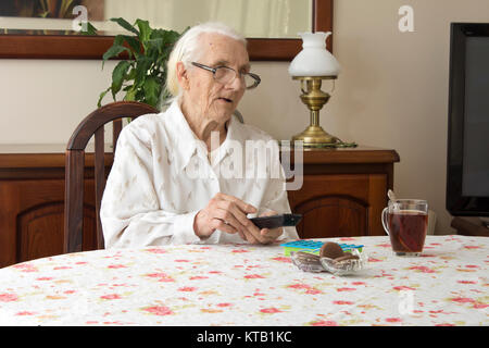 Alte Frau am Tisch mit Fernbedienung für den fernseher. Die alte Frau schaltet die fernbedienung des fernsehers ein. Stockfoto