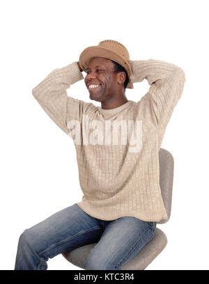 Eine sehr glückliche junge schöne afrikanische amerikanische Mann sitzt auf einem Stuhl mit Hut, seine Hände hinter dem Kopf, auf weißem Hintergrund Stockfoto