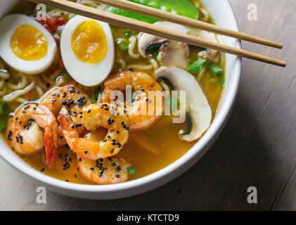 Eine Schüssel mit Ramen mit Ei, Garnelen, Pilzen und Zuckerschoten. Weiße Schüssel, Essstäbchen und Holz Tisch. Stockfoto