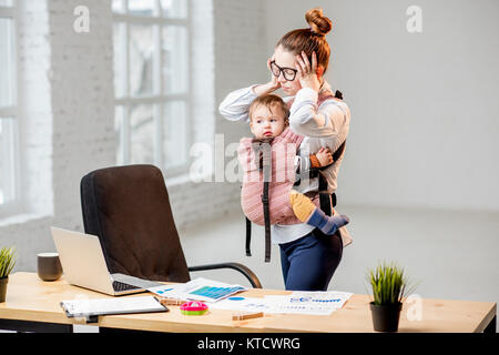 Erschöpft Geschäftsfrau halten ihren Kopf stehend, mit ihrem Baby Sohn im Büro während der Arbeit Stockfoto