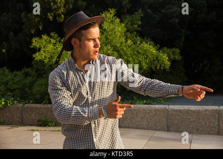 Stark und muskulös junger Mann auf der Straße. Er trägt ein T-Shirt mit braunen Streifen. Das Tragen einer braunen Hut Simulation West Ästhetik. Stockfoto