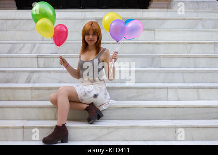 Weiße Mädchen mit orange Haare sitzen auf einem weißen Treppe halten bunte Luftballons mit ihren Händen an einem bewölkten Tag. Stockfoto