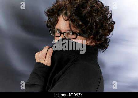 Frau mittleren Alters mit dem lockigen braunen Haar zu tragen schwarze Brille und ihr Gesicht mit ihrem Kragen Stockfoto