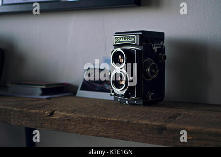 Ein klassischer Vintage Rolleicord Kamera ordentlich positioniert auf einem Holzregal mit der Sonne Auslaufen durch ein großes Fenster mit Fotos auf der Rückseite. Stockfoto