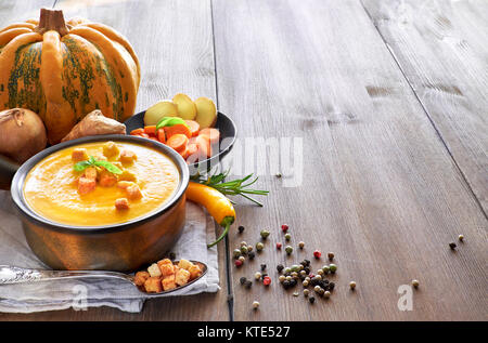 Kürbis, Karotten und Ingwer creme Suppe in Keramik Topf auf dem Holztisch, gewürzt mit Croutons und Basilicum. Zutaten: Kürbis, Karotten, Ingwer, Stockfoto