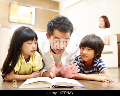 Asiatische Vater und zwei Kinder liegen auf der Vorderseite auf dem Boden lesen Buch, während Mutter aufpassen im Hintergrund. Stockfoto