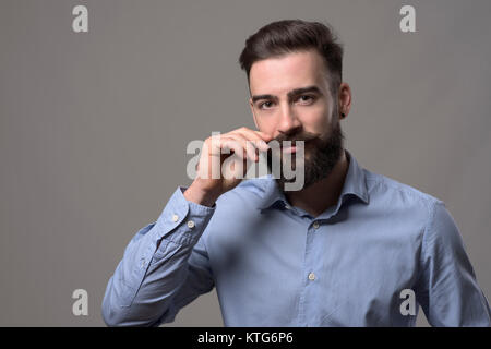 Moody Porträt der jungen stilvolle bärtiger Mann twirling Schnurrbart und Kamera über Grau studio Hintergrund mit Copyspace. Stockfoto