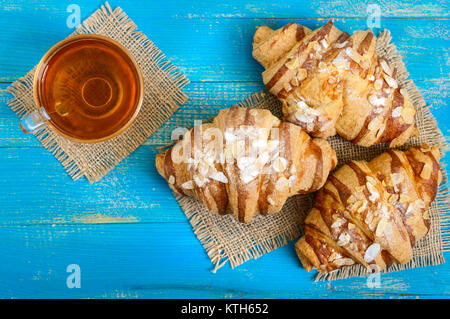 Frisch gebackene Croissants mit Mandelflocken, Tasse Tee auf einem blauen Hintergrund aus Holz. Französisches Gebäck. Frühstück. Stockfoto