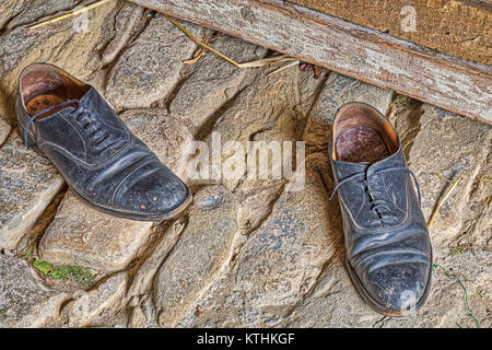 Beat Schwarz Leder Schuhe von alten Bauern auf alten Stein Straße in der Nähe von Grunge Holzbank links Stockfoto