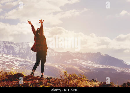 Gerne Frau steht mit erhobenen Händen auf dem Hintergrund der Sonnenuntergang in der Bergkette Stockfoto