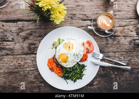 Frühstück mit Spiegelei, Lachs, Avocado, gebackene Tomaten, Toast und Grüns. Tasse aroma Cappuccino auf Holz- strukturierten Hintergrund, flach Stockfoto