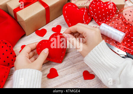 Valentinstag Thema. Arbeitsplatz für die Vorbereitung handgefertigte Dekorationen. Blick von oben auf die weiblichen Händen fühlte Herzen nähen. Verpackte Geschenke, Werkzeuge schäbigen Holztisch. Stockfoto