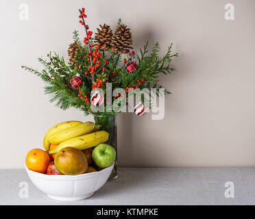 Beeren Tannenzapfen und Frische winter Grün wie ein Blumenstrauß auf einem weißen Tisch vor einer Wand, schöne Weihnachten Dekoration mit Obst in thw Vase Stockfoto