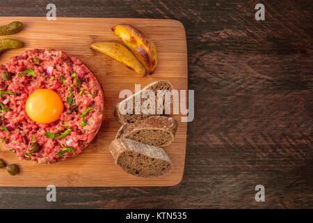 Ein overhead Nahaufnahme eines Steak tartare mit einem rohen Eigelb, Cornichons, Kapern, Roggenbrot, Bratkartoffeln, geschossen von oben auf einen dunklen rustikalen Text Stockfoto