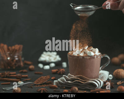 Hand bestreut Zimtpulver auf Glas Becher mit heißer Schokolade Kakao trinken. Kopieren Sie Platz. Der dunkle Hintergrund. Low Key. Winter Essen und Trinken Konzept. Stockfoto