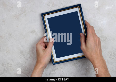 Der alte Mann versucht einen blauen Rahmen aus Holz mit einem Foto an die Wand zu hängen. Konzept Studio gedreht. Bild isoliert auf dunkelblau mit Patch. Stockfoto