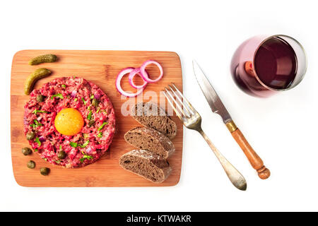 Ein Foto von einem Steak Tartar mit einem rohen Eigelb, Cornichons, Kapern, Roggenbrot, lila Zwiebeln und ein Glas Wein, geschossen von oben auf weißem Hintergrund Stockfoto