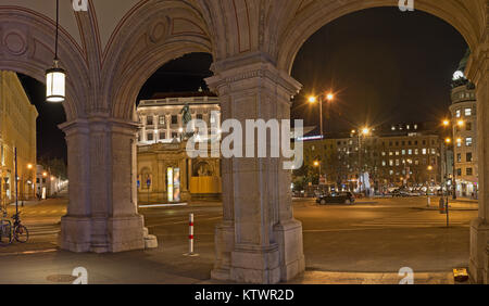 Blick auf den beleuchteten Albertina Square bei Nacht durch Bögen auf Säulen der Staatsoper in Wien. Stockfoto