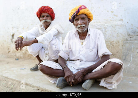 Zwei ältere Indische Männer in Weiß, das Tragen von Turbanen und großen Schnurrbart, auf einem Bürgersteig in einem Dorf in der Nähe von Pushkar, Rajasthan, Indien sitzen. Stockfoto