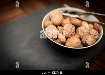 Eine Schüssel gemischten ganze Nüsse in der Schale, einschließlich Walnüsse, Haselnüsse, Mandeln und Pekannüsse mit einem Nussknacker Stockfoto