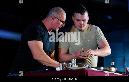 Ein US-Soldat hilft professionellen Koch Robert Irvine Kochen einer Mahlzeit auf der Bühne während der USO Holiday Tour am Al Dhafra Air Base zum 22. Dezember 2017 in der Nähe von Abu Dhabi, Vereinigte Arabische Emirate. Stockfoto