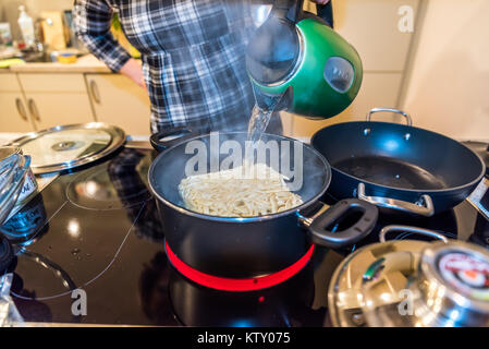 Weibliche hand gießt kochendes Wasser von grün Wasserkocher über Ziegel von gefrorenen Nudeln im Pan-Auflauf auf modernen Elektroherd. Stockfoto