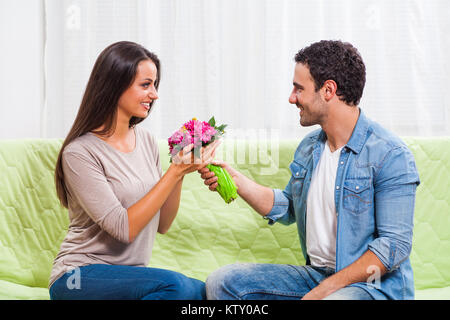 Junges Paar sitzt auf einem Sofa zu Hause. Der Mensch ist mit Blumen zu seiner Frau. Stockfoto
