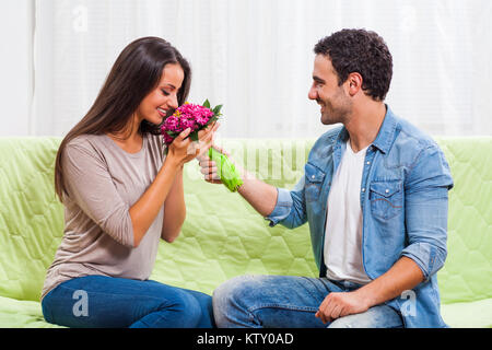 Junges Paar sitzt auf einem Sofa zu Hause. Der Mensch ist mit Blumen zu seiner Frau. Stockfoto