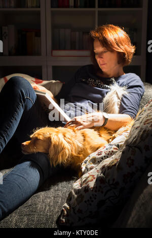 Lesen rothaarige Frau und spaniel Mischling Hund liegen bequem auf dem Sofa vor dem Bücherregal im Wohnzimmer im Licht einer Lampe Stockfoto