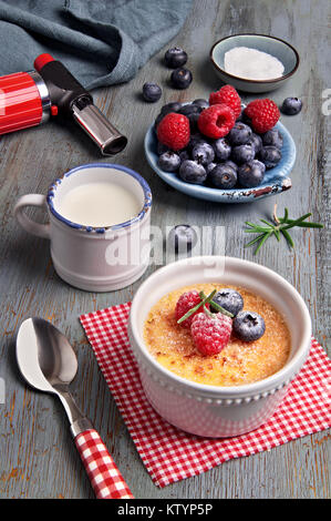 Crème Brûlée mit Himbeeren, Heidelbeeren und Rosmarin in Weiß ramekin mit Zutaten und brennende Fackel auf rustikalen Holztisch. Stockfoto