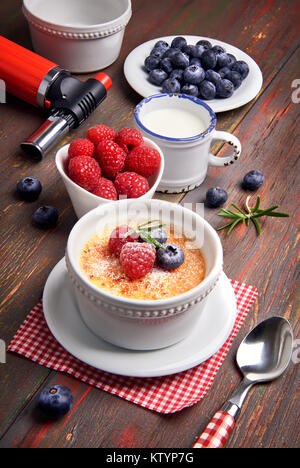 Crème Brûlée mit Himbeeren, Heidelbeeren und Rosmarin in Weiß ramekin mit Zutaten und brennende Fackel auf rustikalen Holztisch. Stockfoto