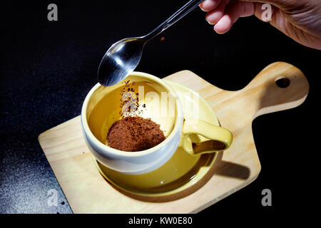 Gießen Sie einen Teelöffel gemahlenen Kaffee in die Tasse. Phasen des Prozesses, eine Tasse heißen Kaffee. Auf schwarzem Hintergrund Stockfoto