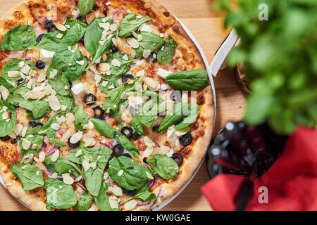 Frische Pizza mit Spinat, Mandeln, Oliven und Parmesan auf hölzernen Tisch im Restaurant. Stockfoto