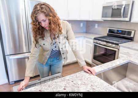 Junge Frau öffnen prüfen Geschirrspüler in modernen Grau, Braun Küche verfügt über Schränke mit Granitoberflächen und fliesenspiegel mit Herd Backofen Stockfoto