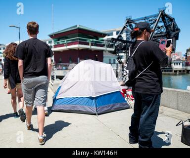 Ein Mann nimmt ein Handy Foto und zwei Leute beiläufig vorbei ein Zelt, das Teil eines Obdachlosen encampment, San Francisco, Kalifornien, 2016. Stockfoto