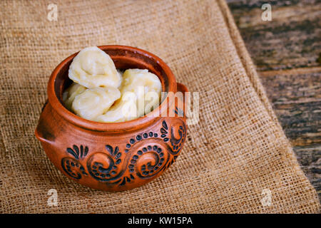 Hausgemachte Teigtaschen pelmeni in einem Tontopf auf einer Leinwand Serviette auf einem hölzernen Hintergrund eingerichtet. Stockfoto