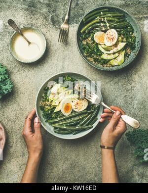 Gesunde vegetarische Frühstück schalen Flachbild-legen. Quinoa, Grünkohl, grüne Bohnen, Avocado, Ei und Tahini dressing Schüsseln in weibliche Hände über konkrete backgroun Stockfoto