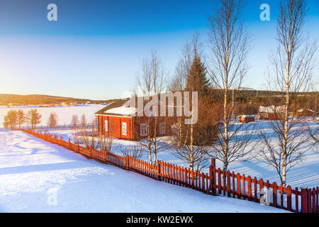 Schöne Winterlandschaft mit Haus am zugefrorenen See in Skandinavien bei Sonnenuntergang Stockfoto