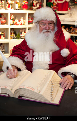 USA Virginia Williamsburg VA Santa Claus Weihnachtsmann Saint Nicolas in seinem Stuhl in seiner Werkstatt, die eine Liste in seinem Buch sitzen Stockfoto