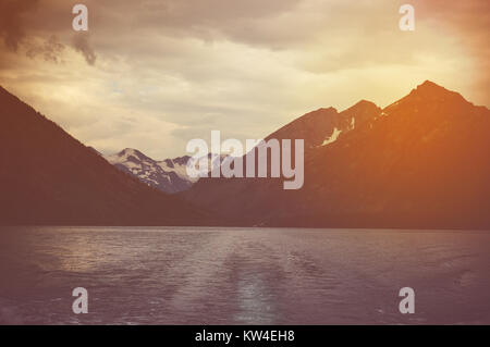 Düstere Landschaft mit dunklen schwarzen See und hohen Klippen mit schneebedeckten Gipfeln im Hintergrund. Den Sonnenuntergang am See, die Sonne hinter den Bergen Stockfoto