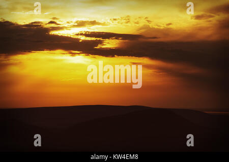 Sonnenuntergang mit Sonnenstrahlen. Hell orange und gelb Strahlen der Sonne leuchten hinter den Wolken auf einem orangefarbenen Himmel über die Hügel Stockfoto