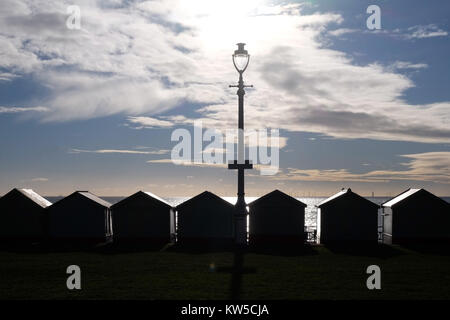 Eine Linie von 7 Umkleidekabinen und eine Straßenlaterne schwarz silhoutted durch die Sonne direkt auf die Kamera über einen dramatischen blau-weißen Himmel, das Meer ist Stockfoto