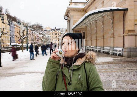 Junge Frau trinkt von Tasse mit therapeutischen Mineralwasser bei einer natürlichen heißen Quelle in Karlsbad im Winter, Tschechische Republik Stockfoto